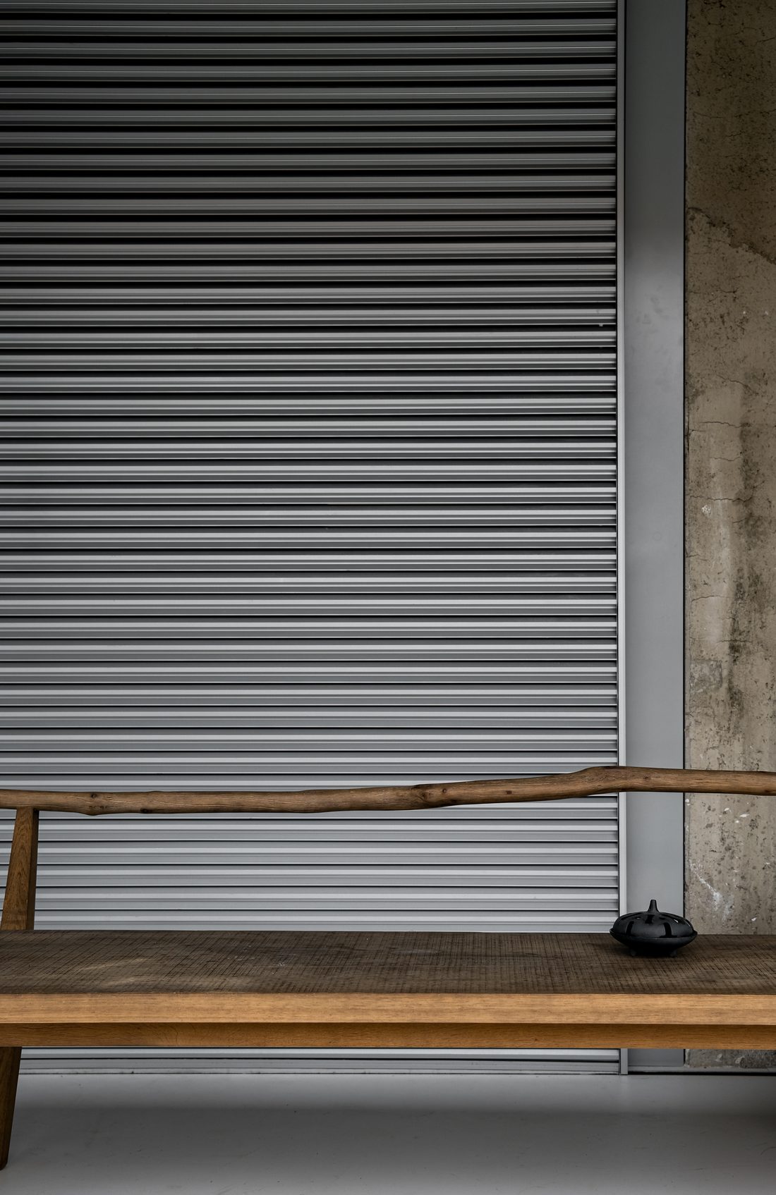 A timber daybed against a metal louvred shutter, with a cast-iron incense burner