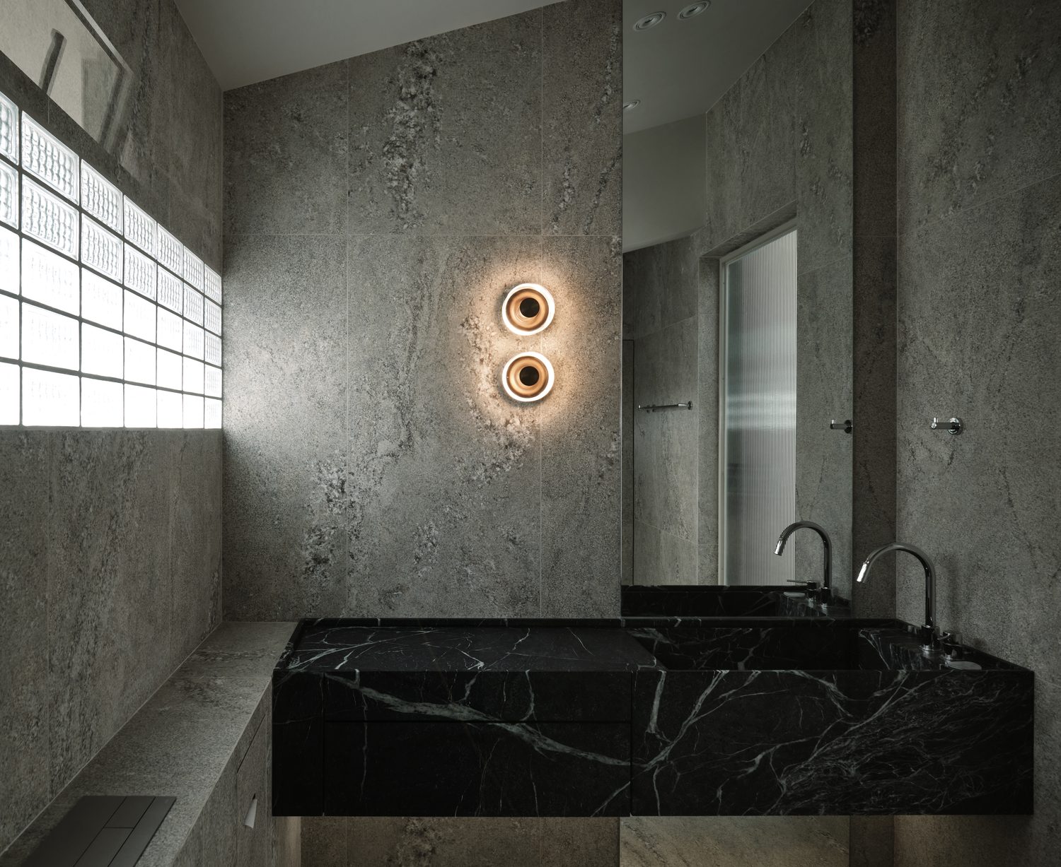 The master bathroom — grey stone and a black-marble twin vanity, a glass-block window, paired ring sconce