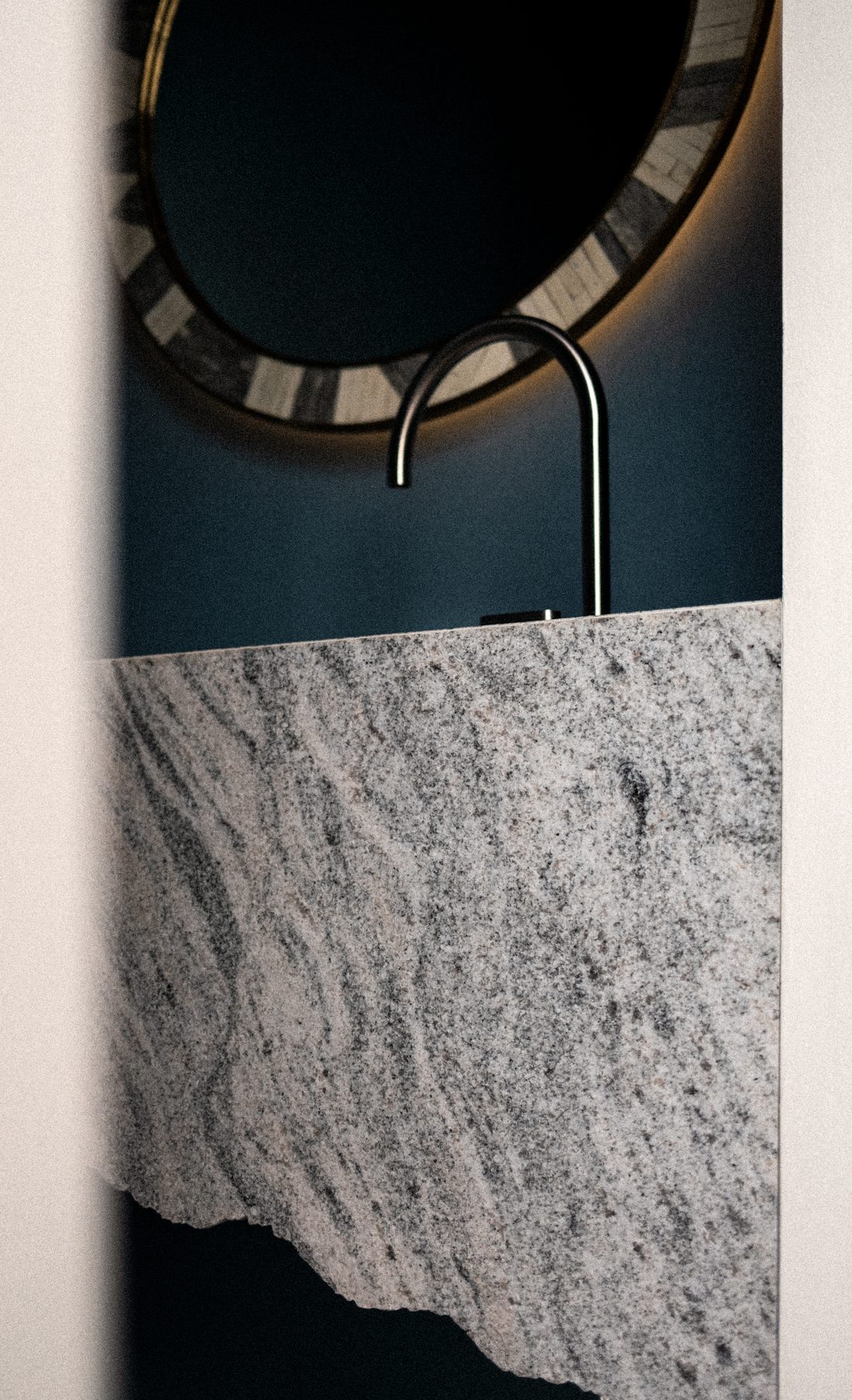 A powder room — a backlit round mirror above a raw granite basin on deep teal