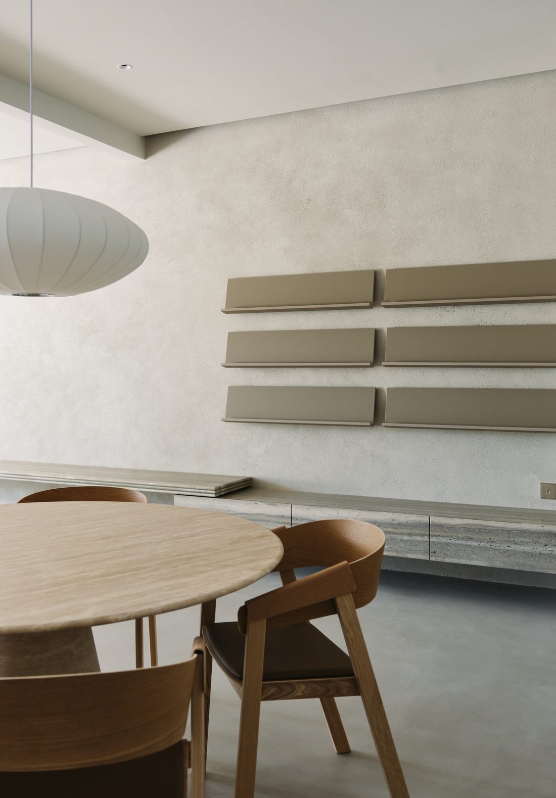 The dining room — a round oak-and-travertine table below a paper pendant, taupe wall shelves beyond