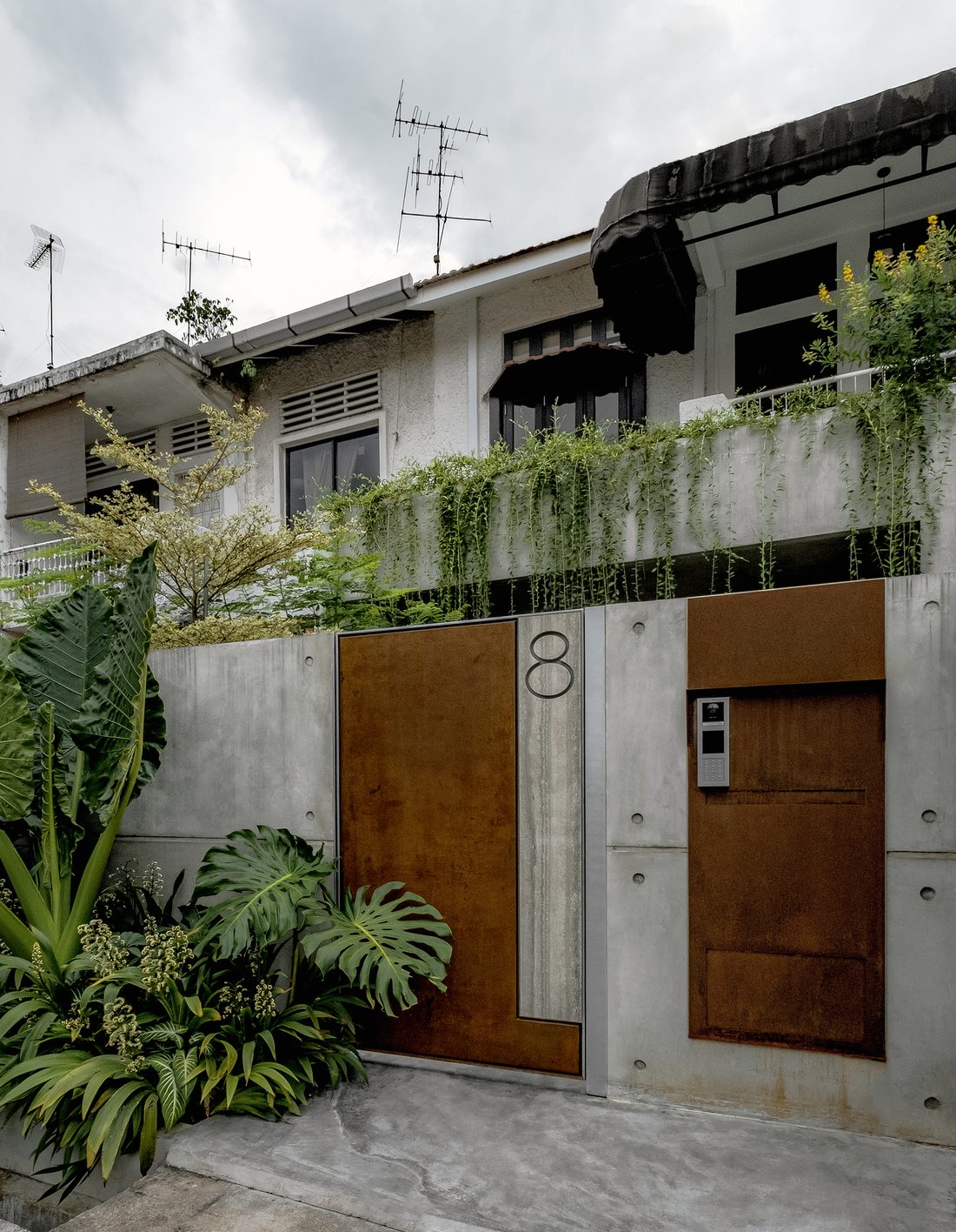 The street facade — board-formed concrete and weathering-steel gates marked no. 8, wrapped in tropical planting