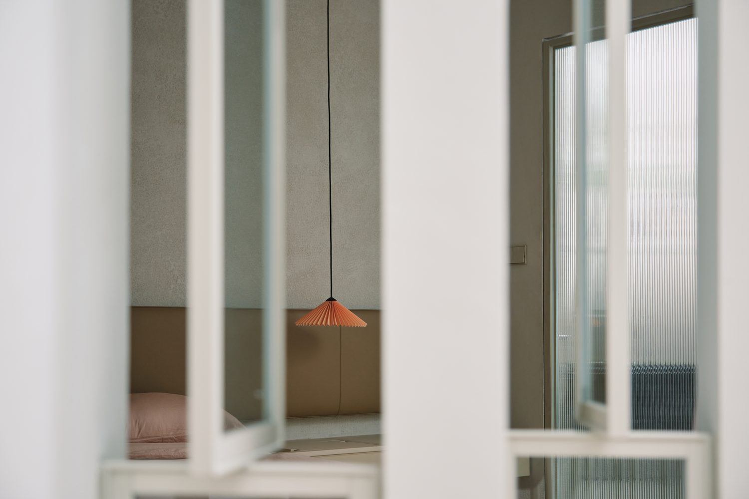 A bedroom glimpsed through a white grid screen, a coral pleated pendant within